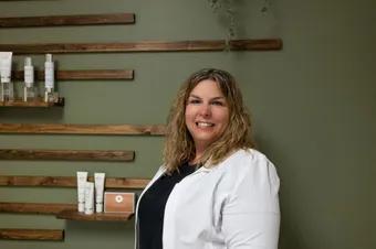 A woman in a white coat is standing in front of a shelf with cosmetics on it.
