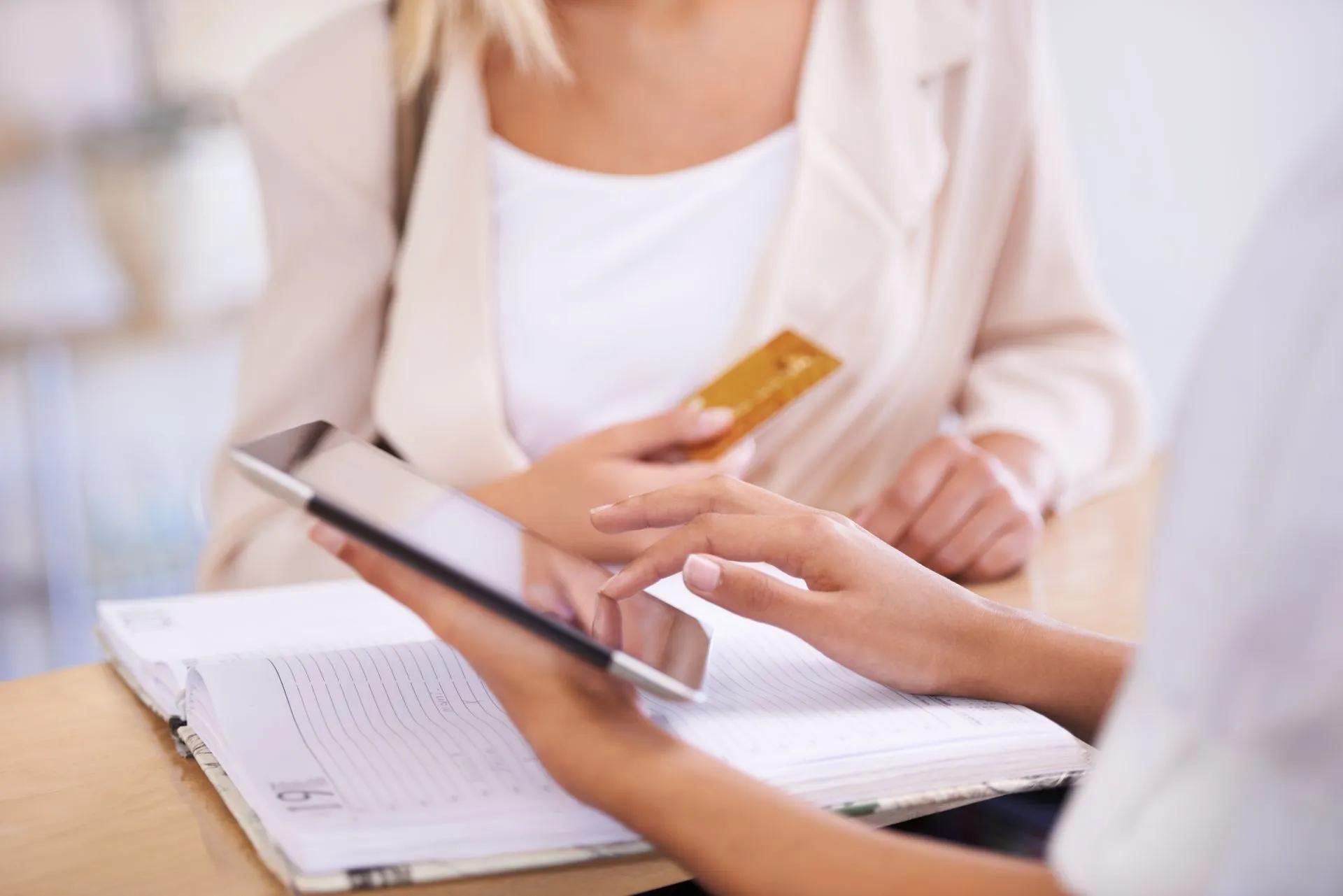 Person handing a gold credit card to another person, who is holding a tablet and a document.